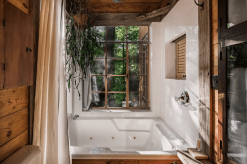 A bright bathroom featuring a large tub, wooden accents, and a window with greenery outside.