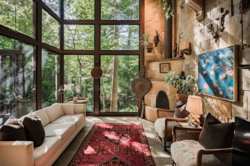 Cozy living room with large windows, a white sofa, a red patterned rug, and greenery visible outside.