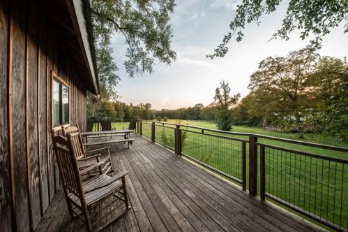 A wooden deck with rocking chairs overlooks a lush green field and trees under a soft evening sky.