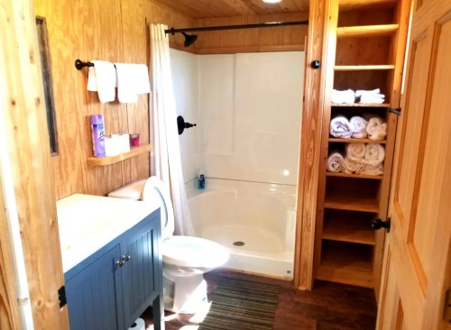 A cozy bathroom featuring a shower, toilet, sink with blue cabinet, and neatly stacked towels on shelves.