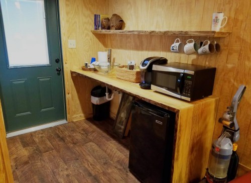 A small kitchen area with a wooden countertop, microwave, coffee maker, and a green door. Shelves hold mugs and utensils.