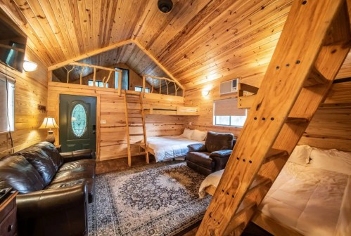Cozy wooden cabin interior with bunk beds, a sofa, and a patterned rug. Warm lighting enhances the rustic atmosphere.