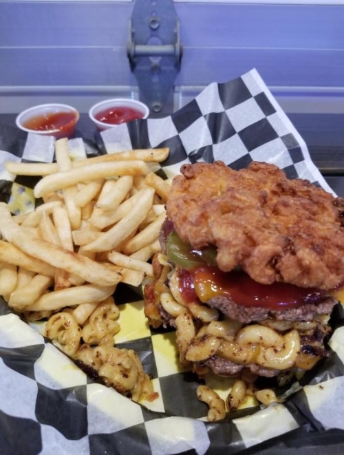 A towering burger with fried chicken, mac and cheese, pickles, and ketchup, served with a side of fries and dipping sauces.