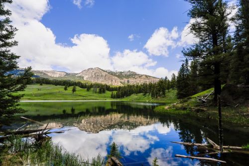 A serene lake reflects mountains and clouds, surrounded by lush greenery and tall trees under a blue sky.