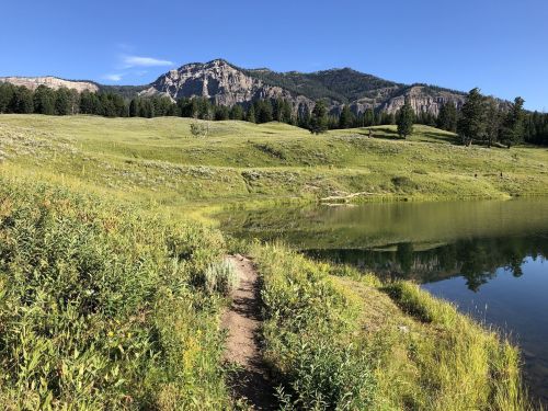 A serene landscape featuring a calm lake, green meadows, and mountains under a clear blue sky.