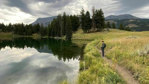 A person stands by a serene lake surrounded by trees and mountains under a cloudy sky.