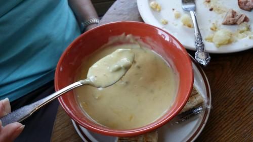 A person holds a spoon above a bowl of creamy soup, with a plate of food and crumbs in the background.