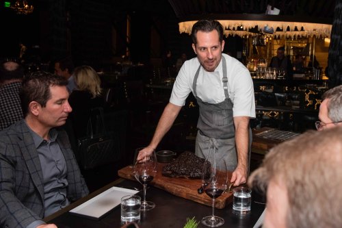 A chef presents a large dish on a wooden board to diners at a restaurant, with wine glasses on the table.