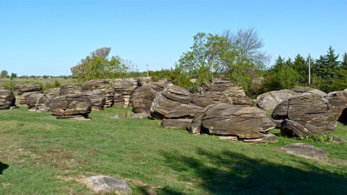 A landscape featuring large, weathered rock formations surrounded by green grass and sparse trees under a clear blue sky.