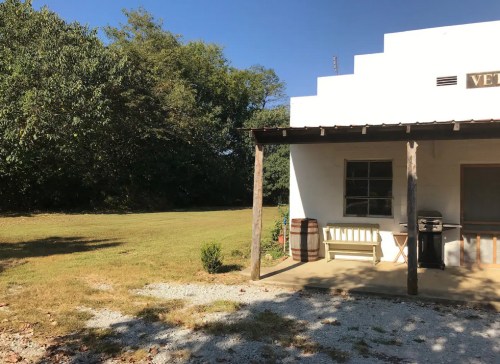 A small white building with a porch, surrounded by green trees and a grassy area under a clear blue sky.