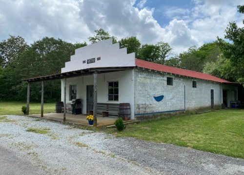 A rustic building with a red roof and white walls, surrounded by greenery and gravel, featuring a porch and barrels.