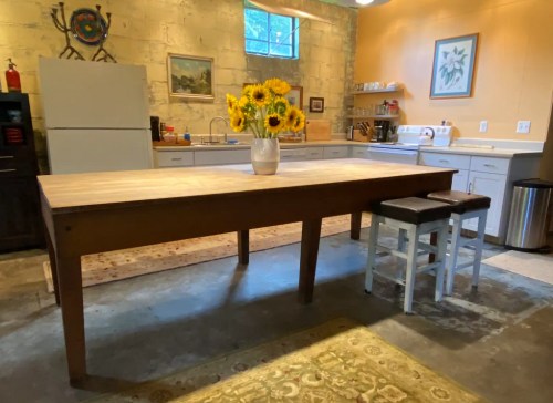 A spacious kitchen with a large wooden table, sunflowers in a vase, and modern appliances against a stone wall.