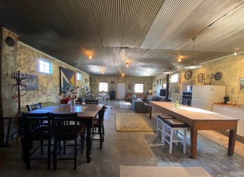 Spacious, rustic interior with dining tables, a living area, and kitchen appliances under a corrugated metal ceiling.