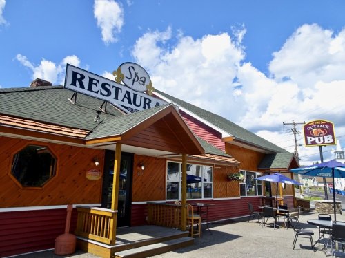 Exterior of a restaurant with a sign reading "Outback Pub," featuring outdoor seating and a blue sky with clouds.
