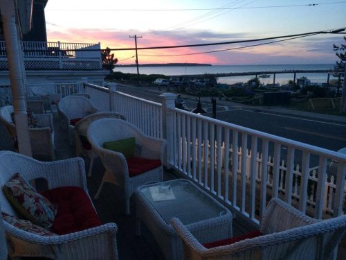 A cozy porch with wicker furniture overlooks a serene sunset by the water, with boats in the distance.