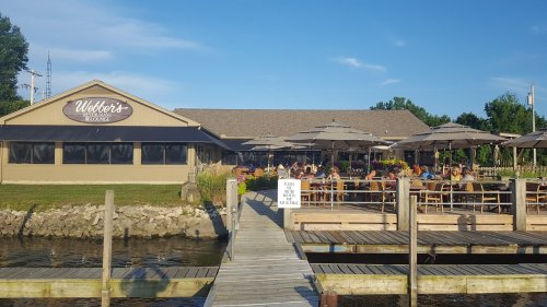 A waterfront restaurant with outdoor seating, umbrellas, and a wooden dock, bustling with diners on a sunny day.