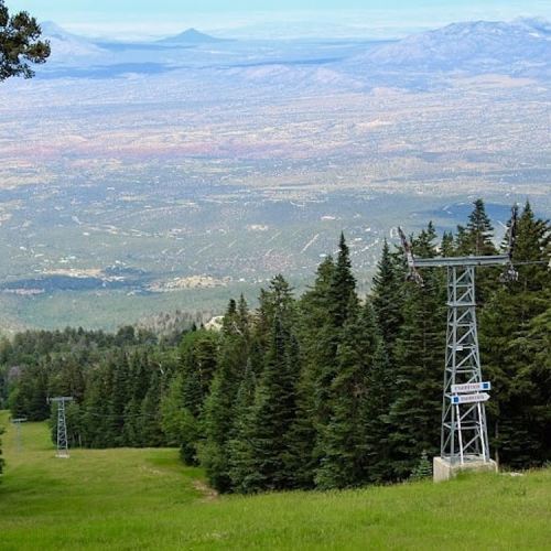 A scenic view from a mountain slope, featuring ski lift towers and lush green trees, with distant mountains in the background.