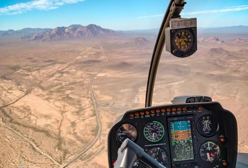 Aerial view from a helicopter cockpit, showcasing desert landscape and distant mountains under a clear blue sky.