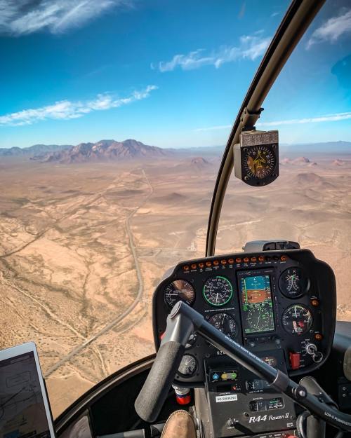 Cockpit view of a helicopter flying over a vast desert landscape with mountains in the distance and a clear blue sky.