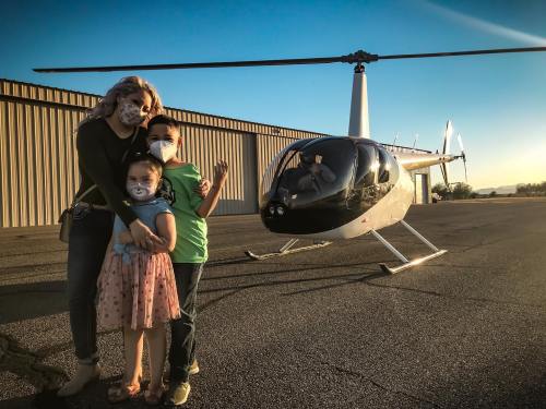 A woman and two children pose with a helicopter on a sunny day, all wearing masks.