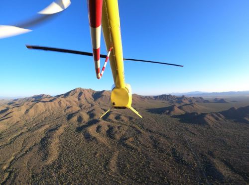 Aerial view from a helicopter flying over rugged mountains and desert landscape under a clear blue sky.