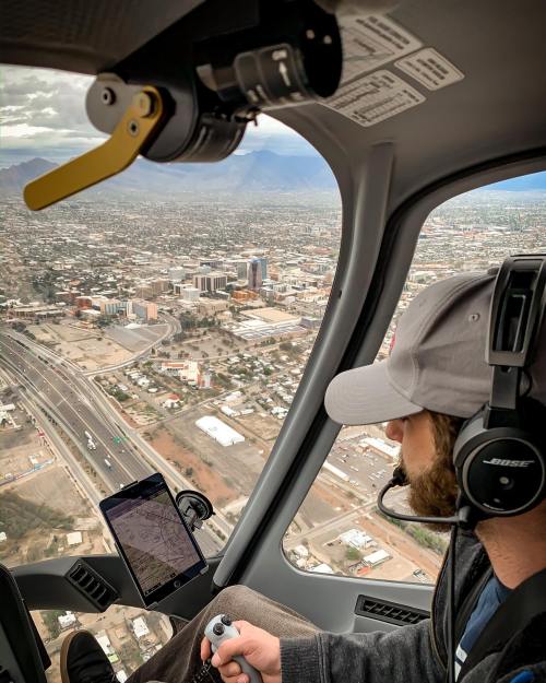 Aerial view from a helicopter showing a pilot using a tablet and controls, with a cityscape below.