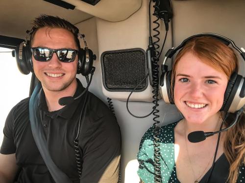 Two smiling individuals wearing headsets inside a helicopter cockpit, ready for a flight.