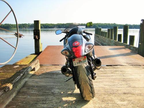 A motorcycle parked on a wooden dock by a calm lake, with trees visible in the background under a clear sky.