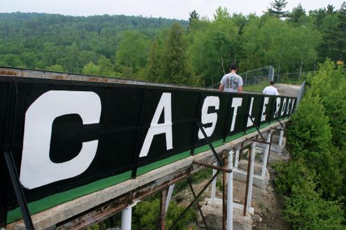 Two people walk along a large sign that reads "CASTLE ROCK," surrounded by lush green trees and hills.