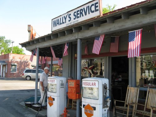 A vintage gas station with American flags, featuring old fuel pumps and a man cleaning outside.