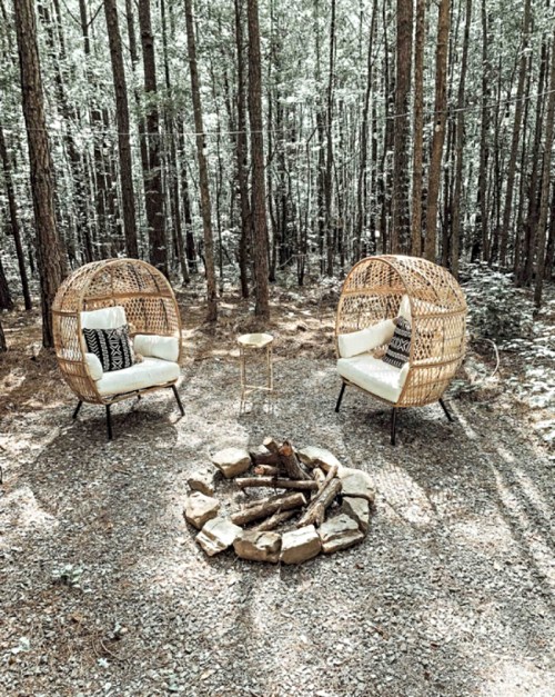 Two cozy wicker chairs with cushions surround a stone fire pit in a serene forest setting.