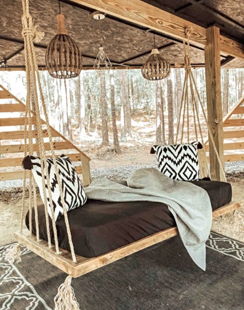 A cozy outdoor swing bed with patterned pillows, draped in a gray blanket, surrounded by trees and rustic decor.