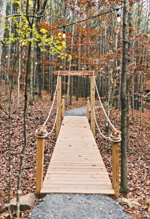 A wooden bridge with rope railings leads through a forest of autumn leaves and trees, illuminated by string lights.