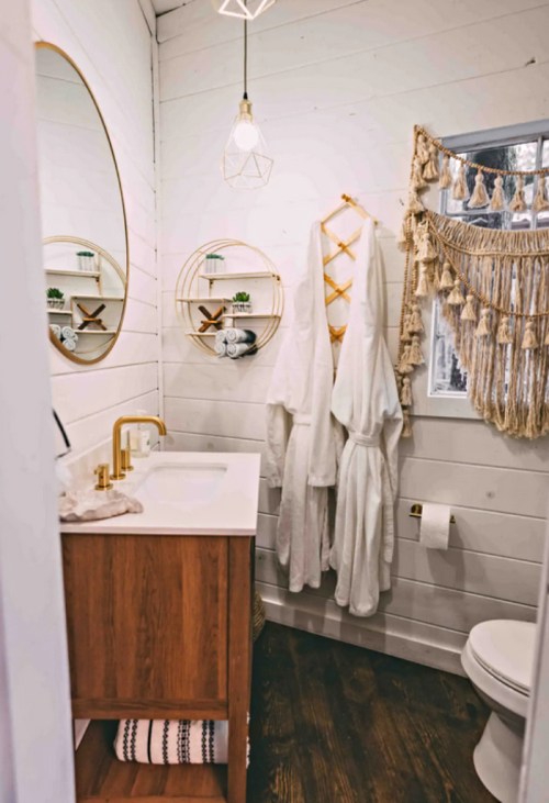 Cozy bathroom featuring a wooden vanity, white robes, decorative shelves, and a stylish mirror against a shiplap wall.