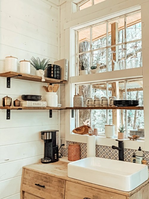 Cozy kitchen corner with shelves, a sink, coffee maker, and decorative items, featuring natural light from large windows.