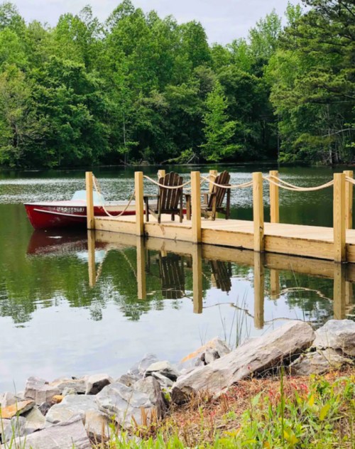 A wooden dock extends over calm water, with a red boat tied up and chairs facing the serene green landscape.