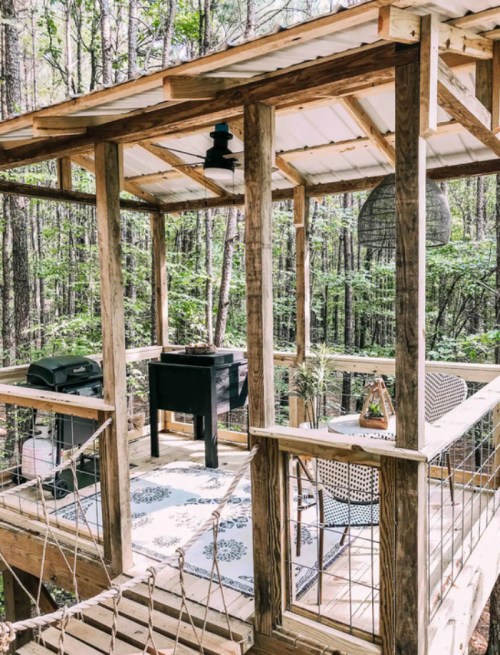 A cozy wooden deck in a forest, featuring a grill, table, and chairs, surrounded by trees and greenery.