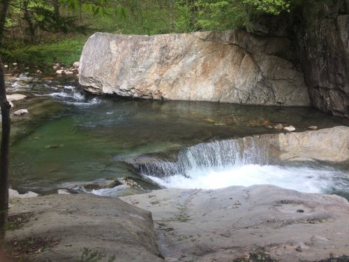 A serene river flows over rocks, creating a small waterfall surrounded by lush greenery and a rocky landscape.
