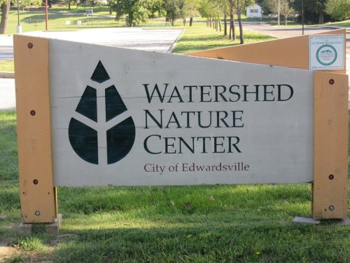 Sign for Watershed Nature Center in Edwardsville, featuring a tree logo and surrounded by green grass.