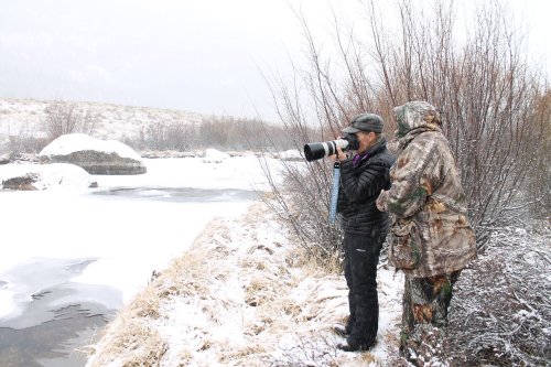 Two people stand by a snowy riverbank, one photographing with a large camera while the other observes.