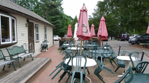 Outdoor seating area with green tables and chairs, pink umbrellas, and a building in the background surrounded by trees.
