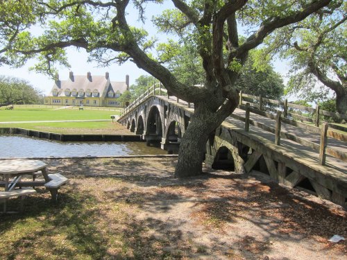 A wooden bridge leads to a large yellow house, surrounded by trees and a calm pond on a sunny day.