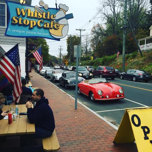 A red sports car parked outside Whistle Stop Cafe, with diners at outdoor tables and American flags lining the street.
