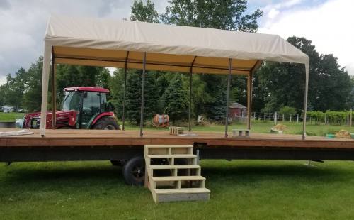 A wooden platform with a tan canopy, steps leading up, and a red tractor in the background on a grassy field.