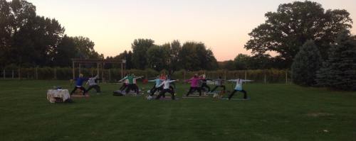 A group of people practicing yoga outdoors on mats during sunset, surrounded by trees and a grassy field.