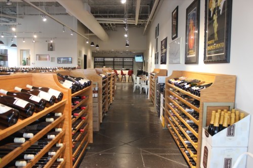 Interior of a wine store with wooden racks filled with bottles, posters on the walls, and a tasting area in the background.
