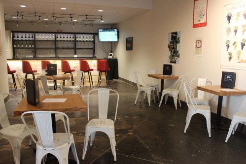 A modern bar area with red stools, empty tables, and a TV displaying a sports game in a well-lit space.