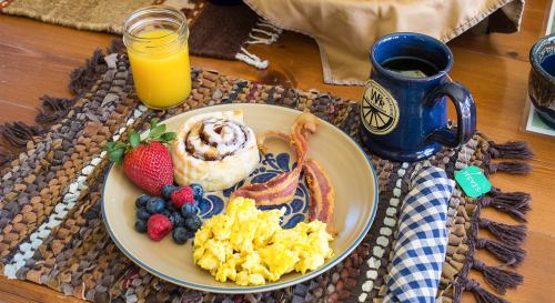 A breakfast plate with scrambled eggs, bacon, a cinnamon roll, strawberries, blueberries, coffee, and orange juice.