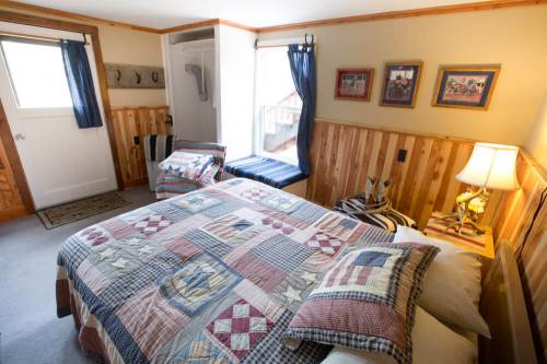 Cozy bedroom with a quilted bed, wooden walls, a window seat, and framed pictures. Soft lighting from a bedside lamp.