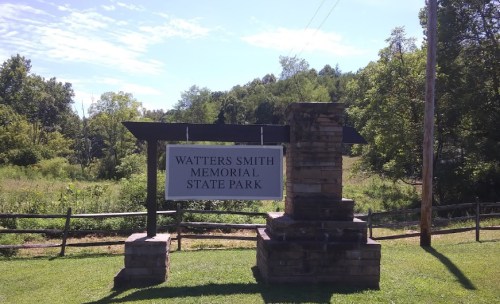 Sign for Watters Smith Memorial State Park, surrounded by greenery and trees under a clear blue sky.
